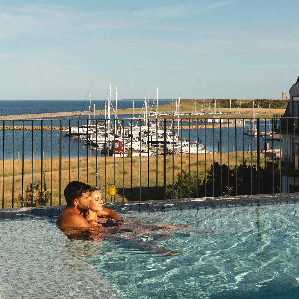 Couple relaxing in Rox Resort's rooftop pool with a cocktail, overlooking Køge marina and the sea.