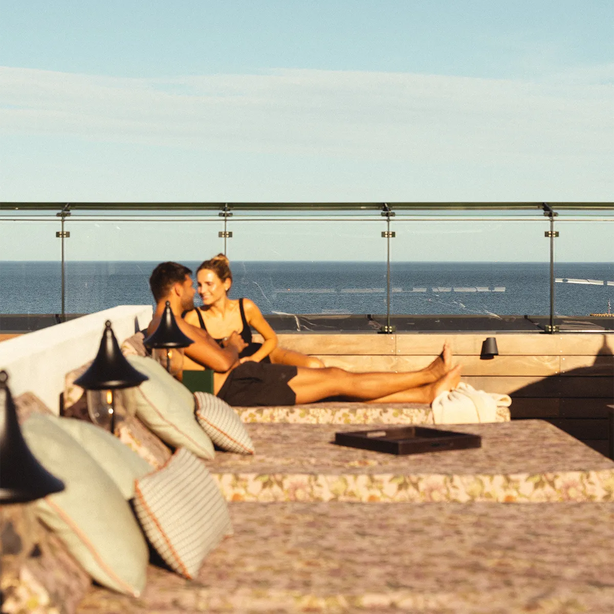 Couple lounging on a cushioned daybed on Rox Resort's rooftop terrace in evening sun, sea in the background.