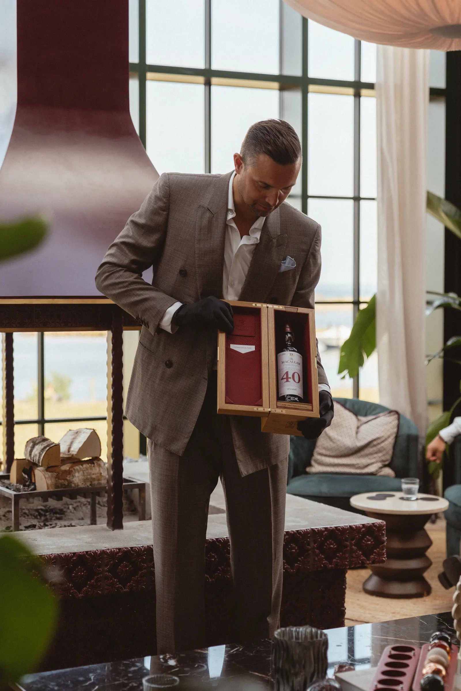 Man in a double-breasted suit and black gloves presenting a bottle of 40-year-old Macallan in its wooden box.