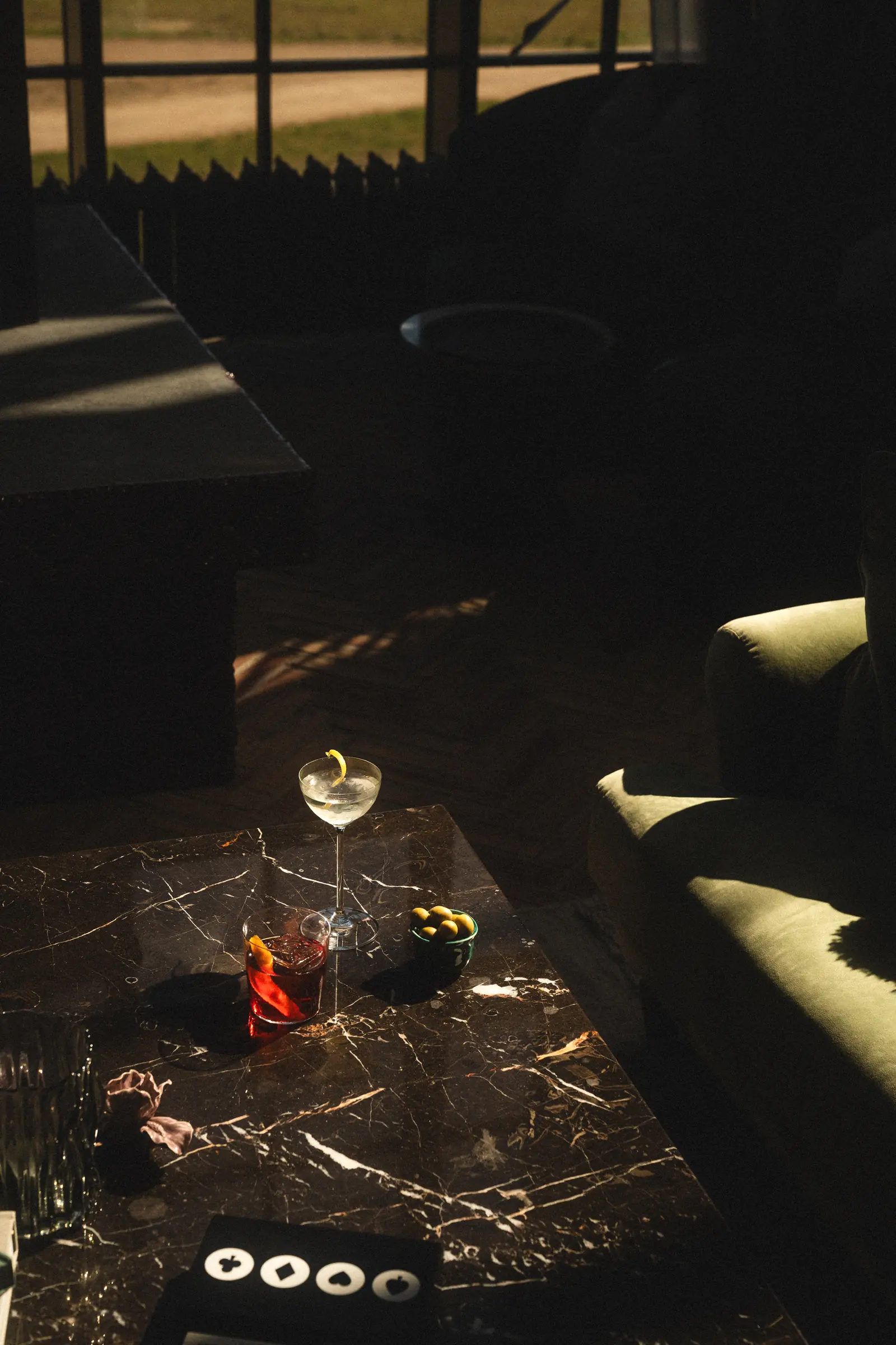 Martini with a lemon twist and a negroni on a dark marble table, with a bowl of olives in dramatic sunlight.