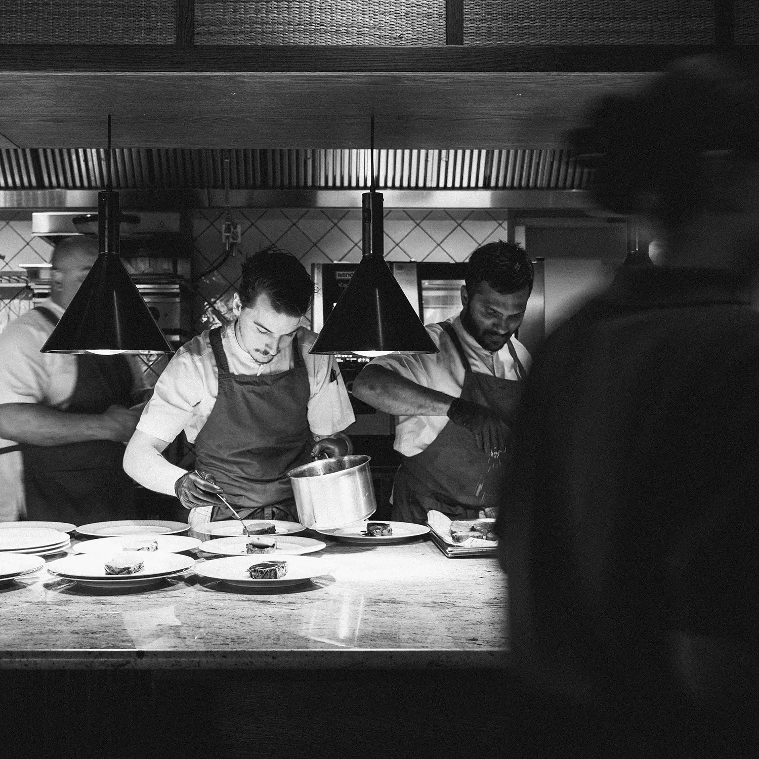 Black-and-white photo of chefs plating dishes at the pass in an open kitchen under two pendant lamps.