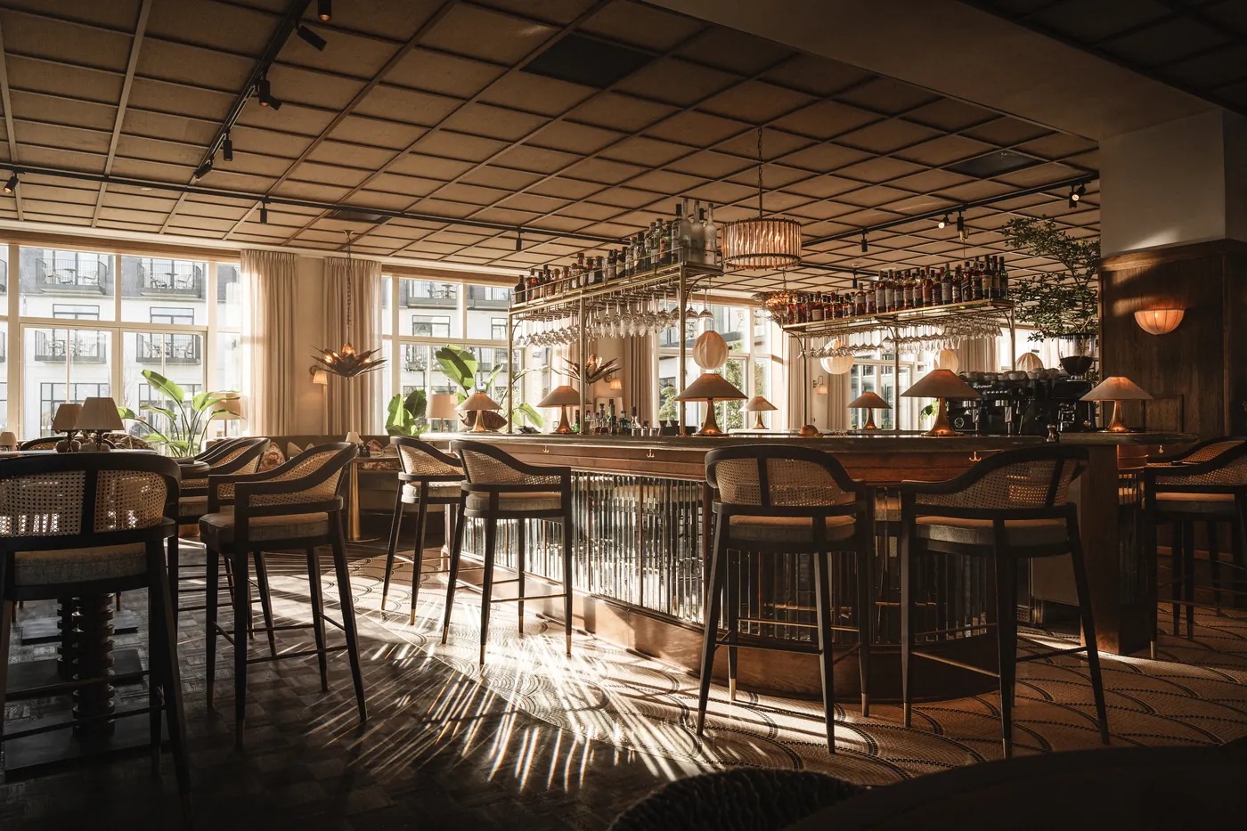 Sunlit bar counter with rattan bar stools, brass shelves of bottles and light patterns across the mosaic floor.