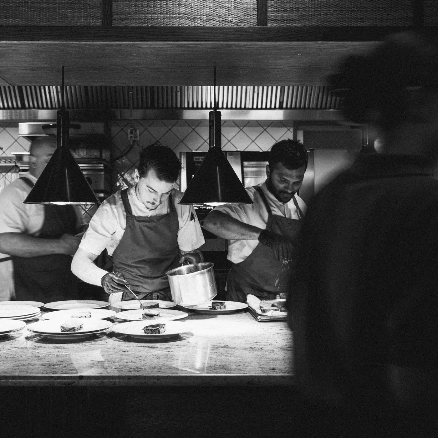 Black-and-white photo of chefs plating dishes at the pass in an open kitchen under two pendant lamps.
