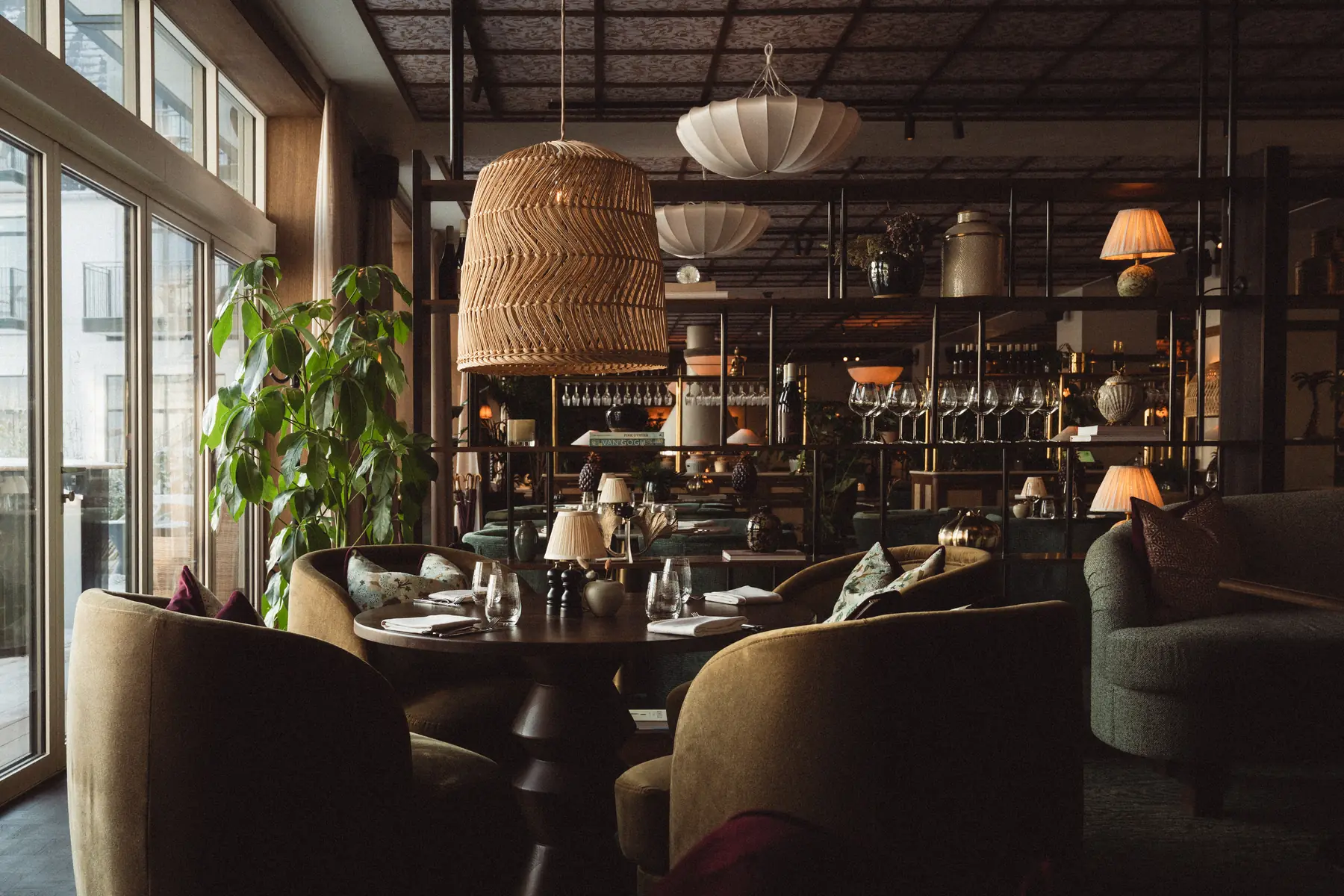 Dimly lit restaurant dining area with a large rattan pendant lamp, round table and olive-green velvet armchairs.