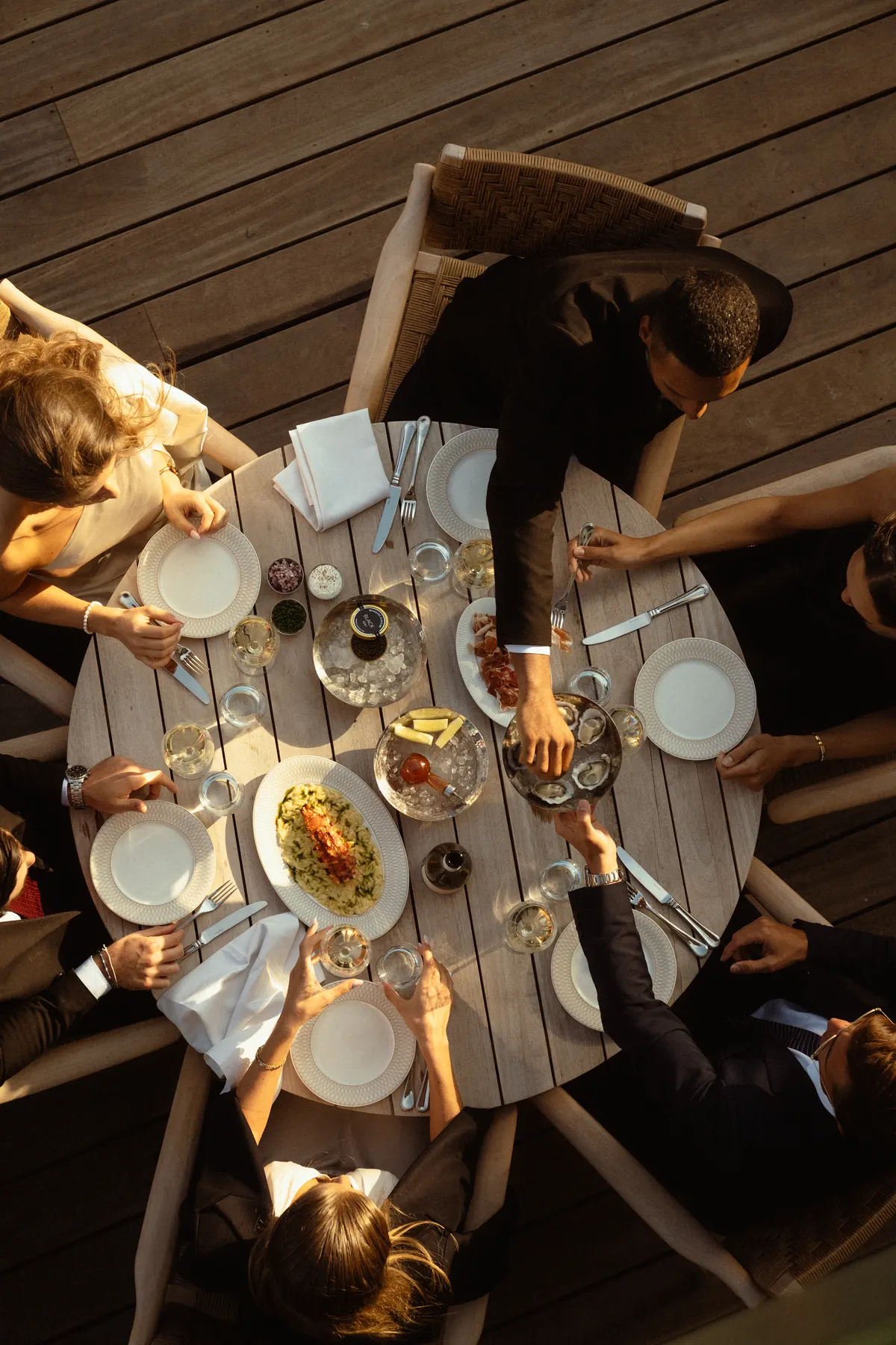 Overhead view of a round table with oysters, caviar on ice, risotto and white wine as a waiter serves guests.