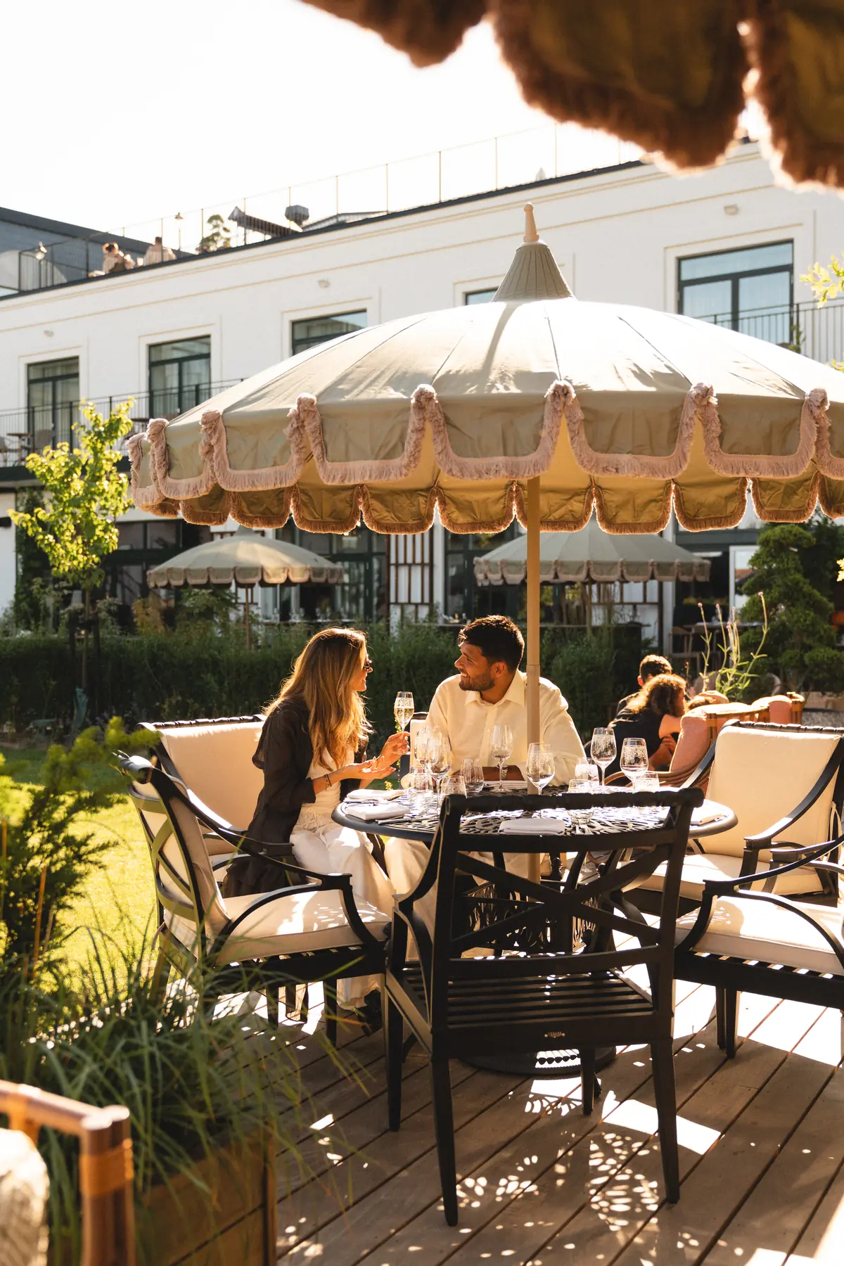 Couple sharing champagne at a garden table under a fringed parasol on a sunny terrace.